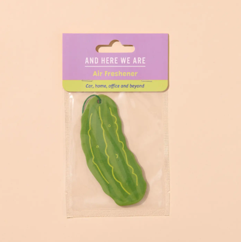 A green pickle-shaped air freshener by And Here We Are in its purple and lime green packaging.