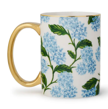 A white porcelain mug by Rifle Paper Co. featuring a wrap-around pattern of light blue hydrangeas and a gold handle.