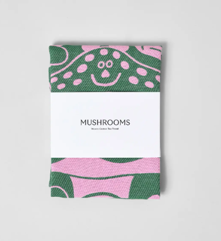 The "Mushrooms" tea towel by Wrap, folded neatly with its paper band, showcasing the unique pink and green color scheme and humorous mushroom illustrations.