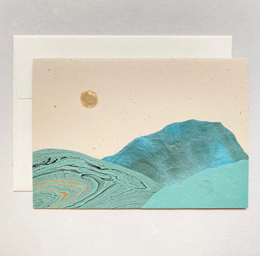 A handmade card featuring a landscape collage with turquoise mountains and a gold moon against a neutral background.