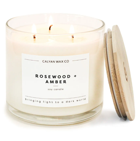 Bring cozy warmth to your home decor with the Rosewood + Amber 3-wick soy candle by Calyan Wax Co., featuring a clear glass tumbler and wooden lid.
