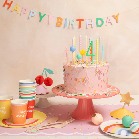 A beautifully styled pink birthday cake topped with colorful Meri Meri balloon candles, surrounded by matching party tableware and a "Happy Birthday" banner.