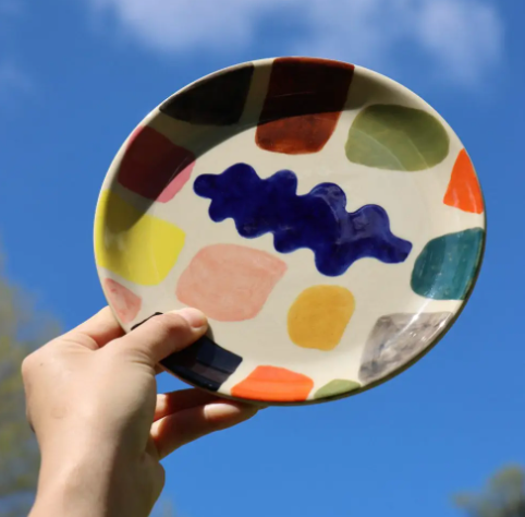 A hand holding up a colourful hand-painted Mottle side plate against a bright blue sky, showcasing its unique abstract palette.