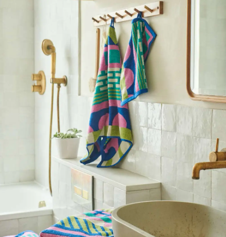 Several colorful patterned towels hanging neatly on wooden hooks in a bright, modern tiled bathroom with gold fixtures.