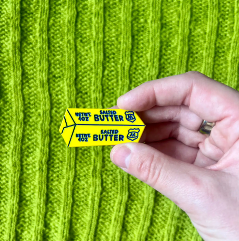 The Boogie Bee Butter Flat Clip held in a hand against a bright green knitted background, showing the yellow clip design with "SALTED BUTTER" graphics.