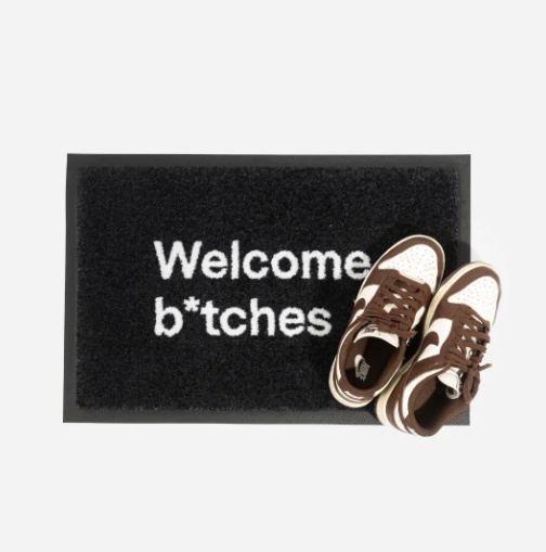 A pair of sneakers partially on the Welcome B*tches doormat.