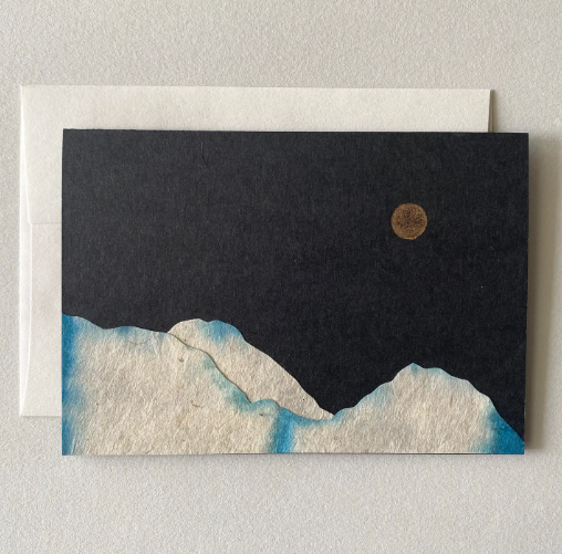 A handmade card featuring a landscape collage with cloud-like mountains and a moon against a dark background.