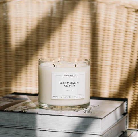 Enjoy a serene, sunlit corner with the lit Oakmoss + Amber 3-wick candle by Calyan Wax Co. resting on stacked hardcover books.