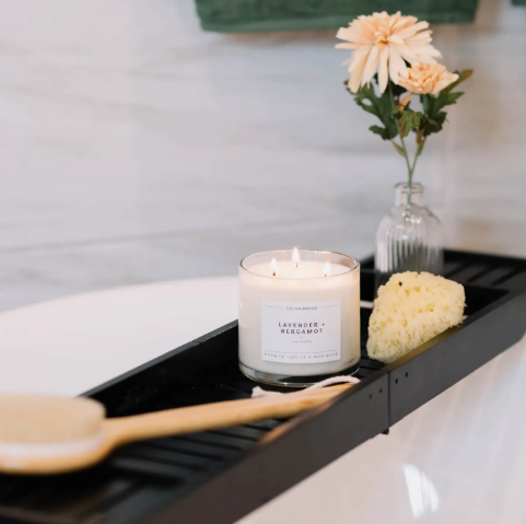 Relax with the lit Lavender + Bergamot 3-wick candle by Calyan Wax Co. displayed on a modern bath caddy beside fresh florals.