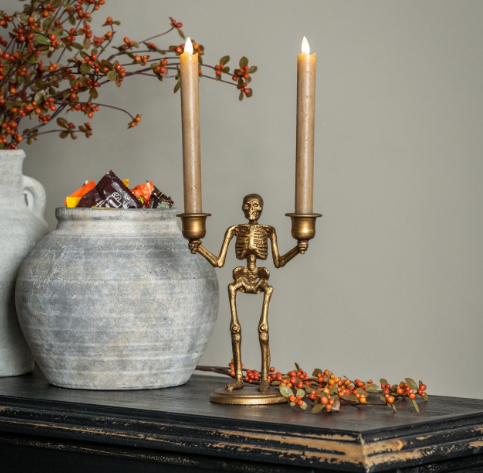 Create a dramatic autumn vignette with the Gold Skeleton Taper Candle Holder by Ragon House, styled with glowing taper candles alongside rustic vessels and berries on a dark console table.