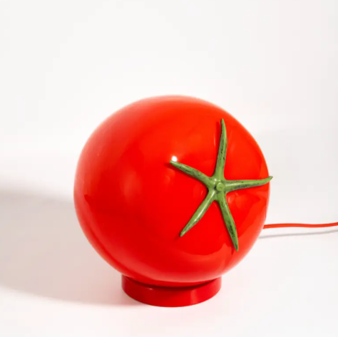 A vibrant red tomato-shaped lamp made of glossy glass with a green stem detail, sitting on a matching red base.