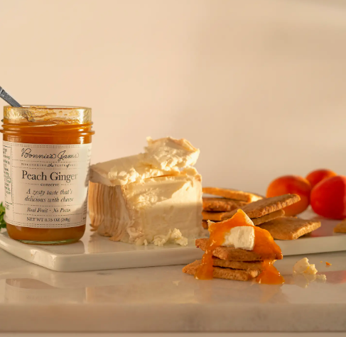 An open jar of Peach Ginger Jam with a spoon inside, ready to be served alongside a fresh slice of cheese.