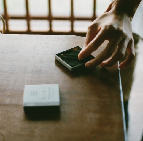 A lit hibi incense match resting safely on its specialized burning pad inside the box by Ameico.