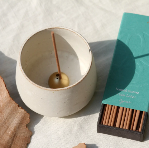 A hand-thrown white ceramic incense bowl with a gold dome holder and burning stick.