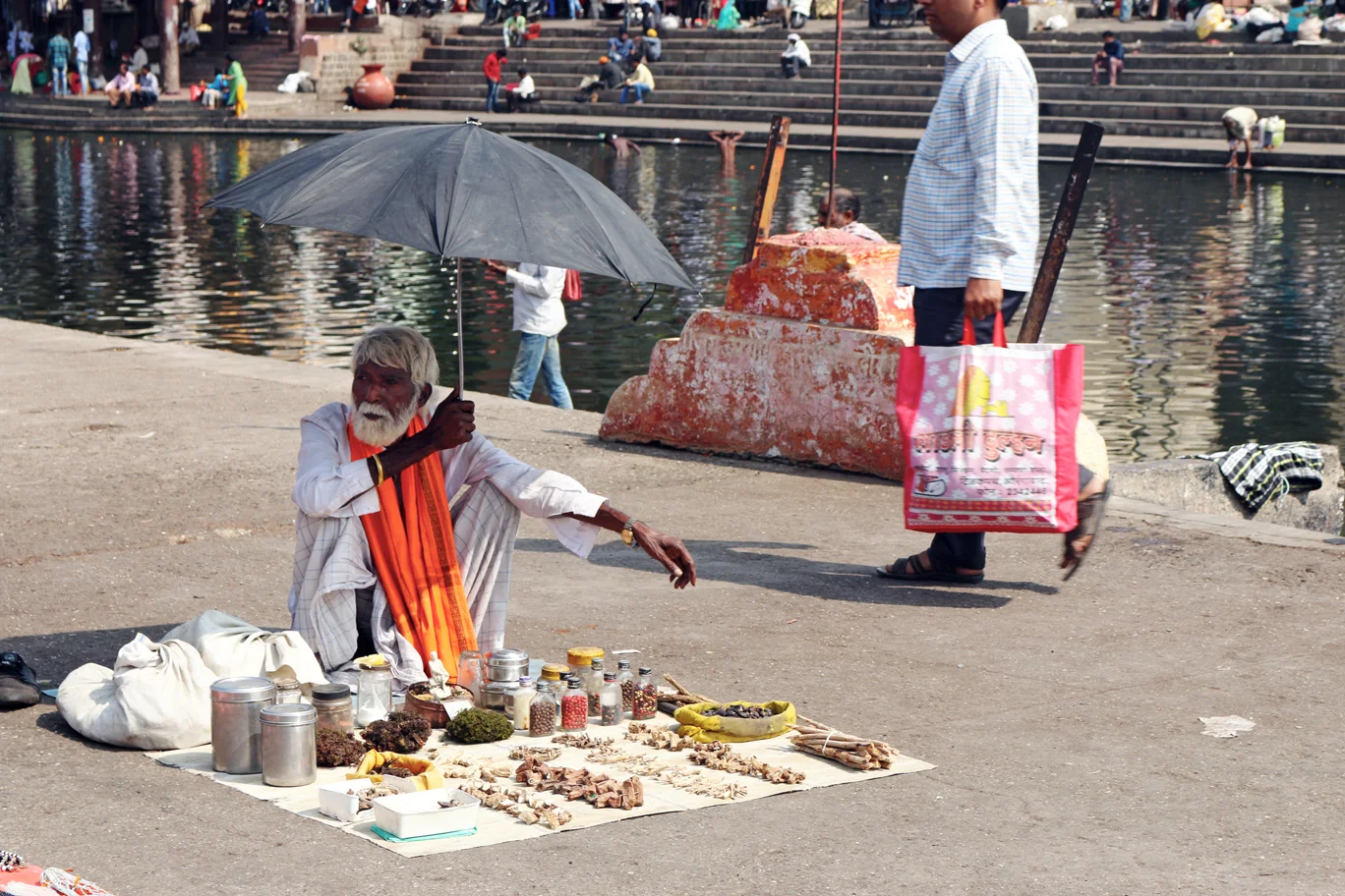 Holy man sells talismans under the shade of an umbrella, while people bathe in the River Ganges