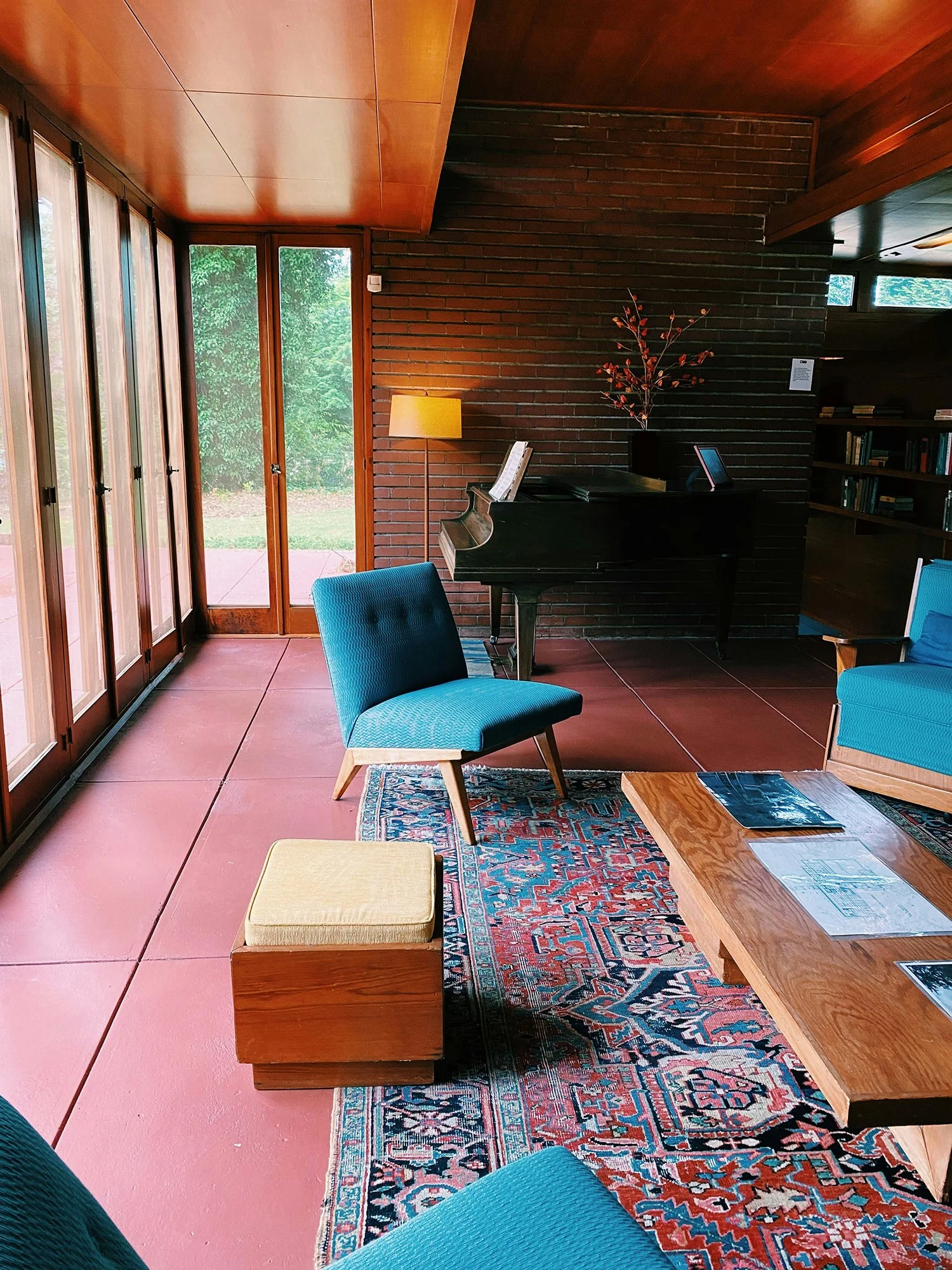 Living room with teal chairs, a wooden coffee table with magazines, a patterned rug, a brick wall, a black piano with sheet music, a floor lamp, and large glass doors leading outside.