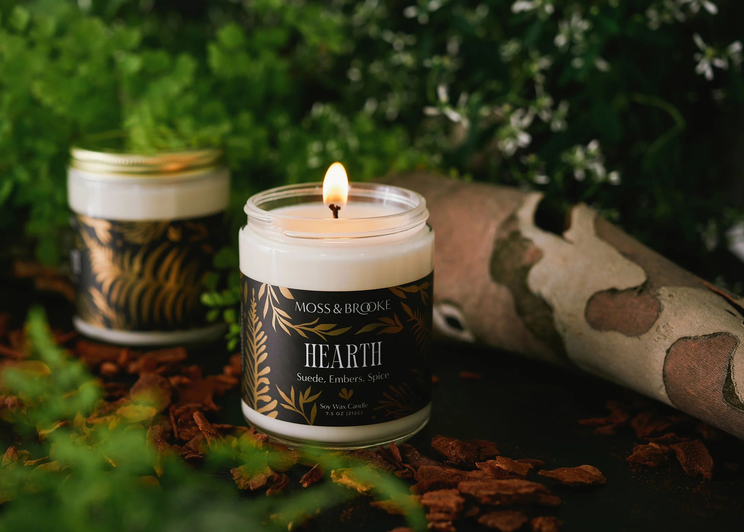 A lit candle surrounded by natural elements like bark, ferns and small white flowers. Photographed by Tina Crespo.