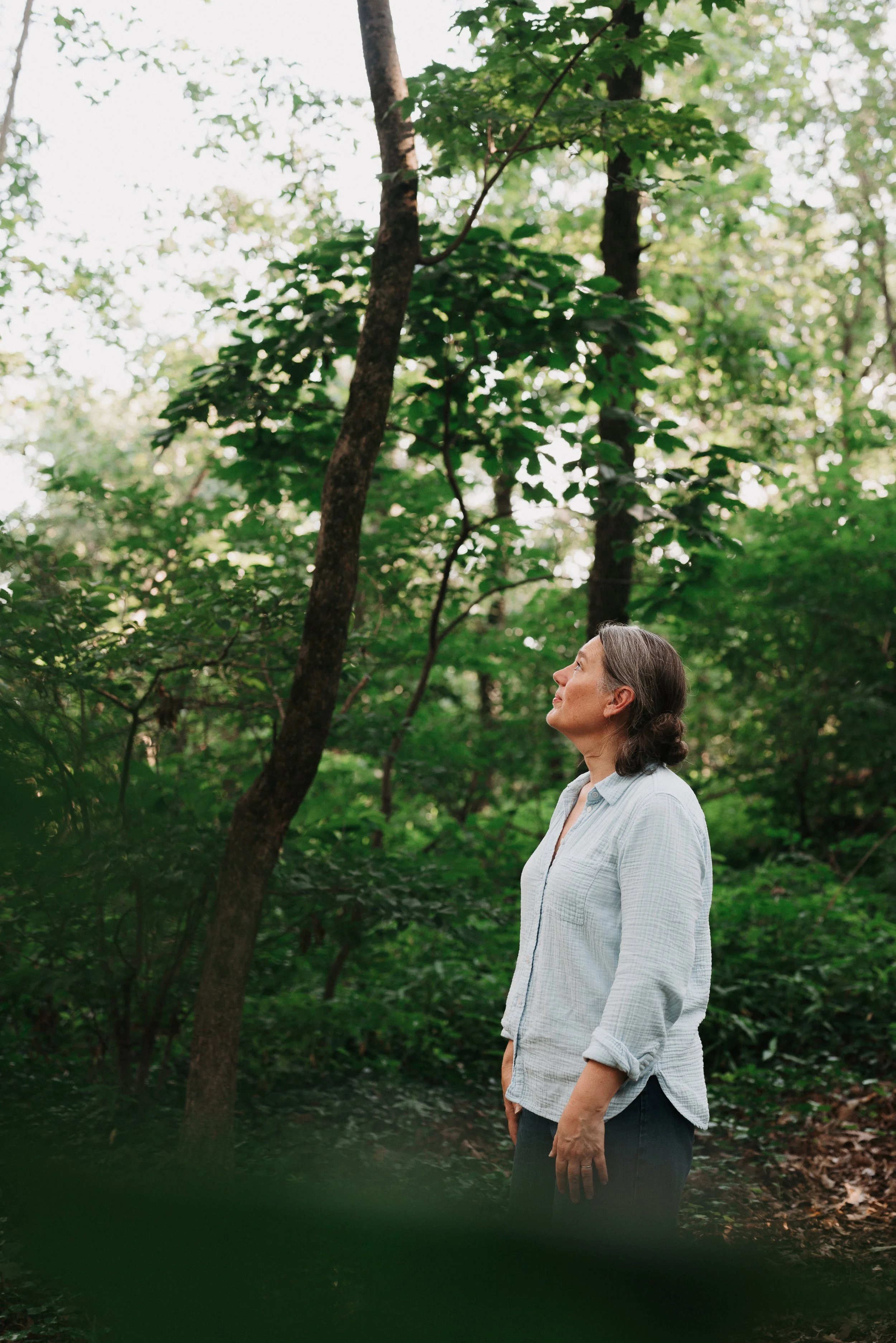 Portrait of a woman in the woods looking up at the trees.