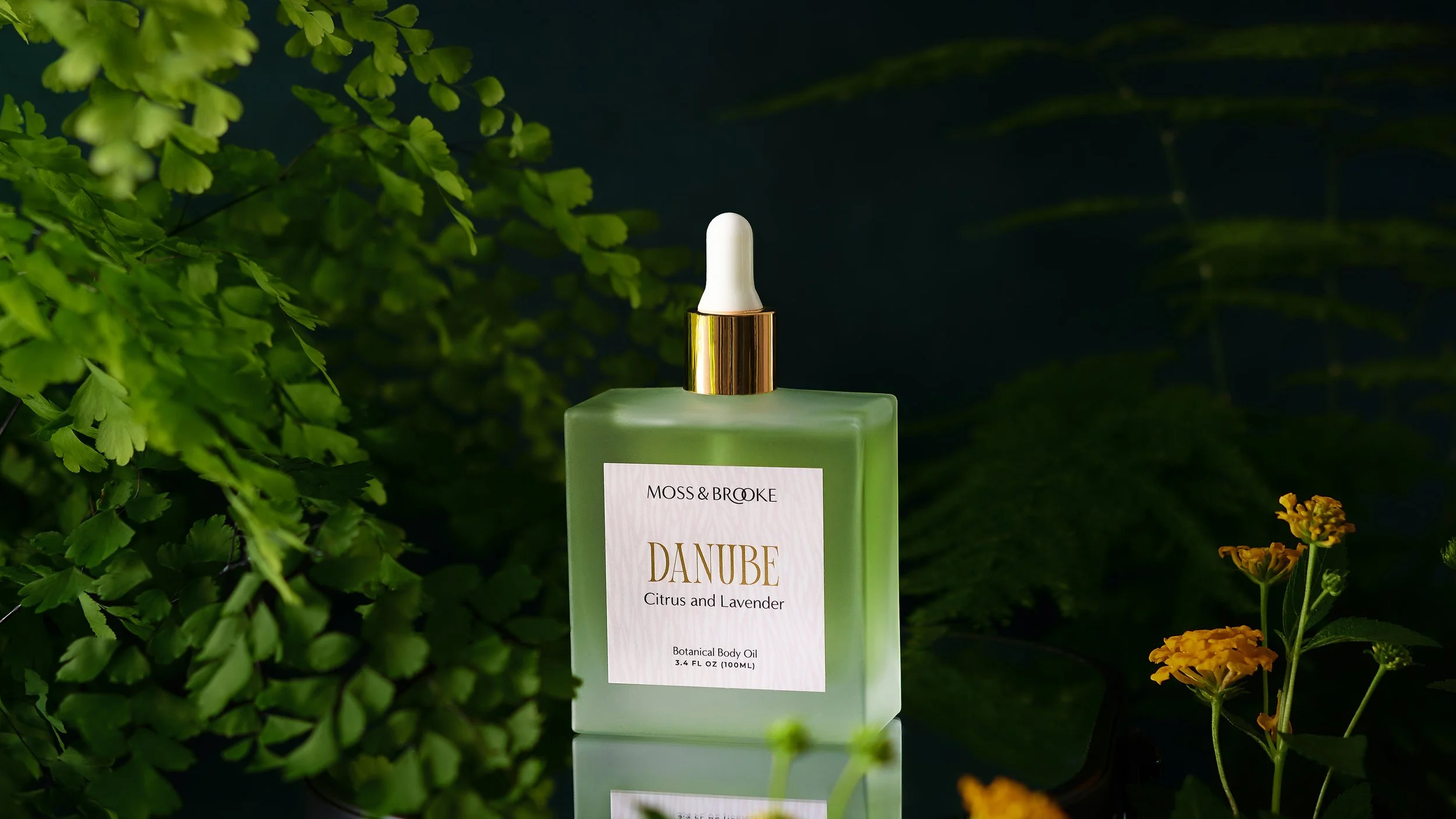 Frosted glass bottle of body oil against a dark background with ferns and marigold flowers surrounding it. Photographed by Tina Crespo.