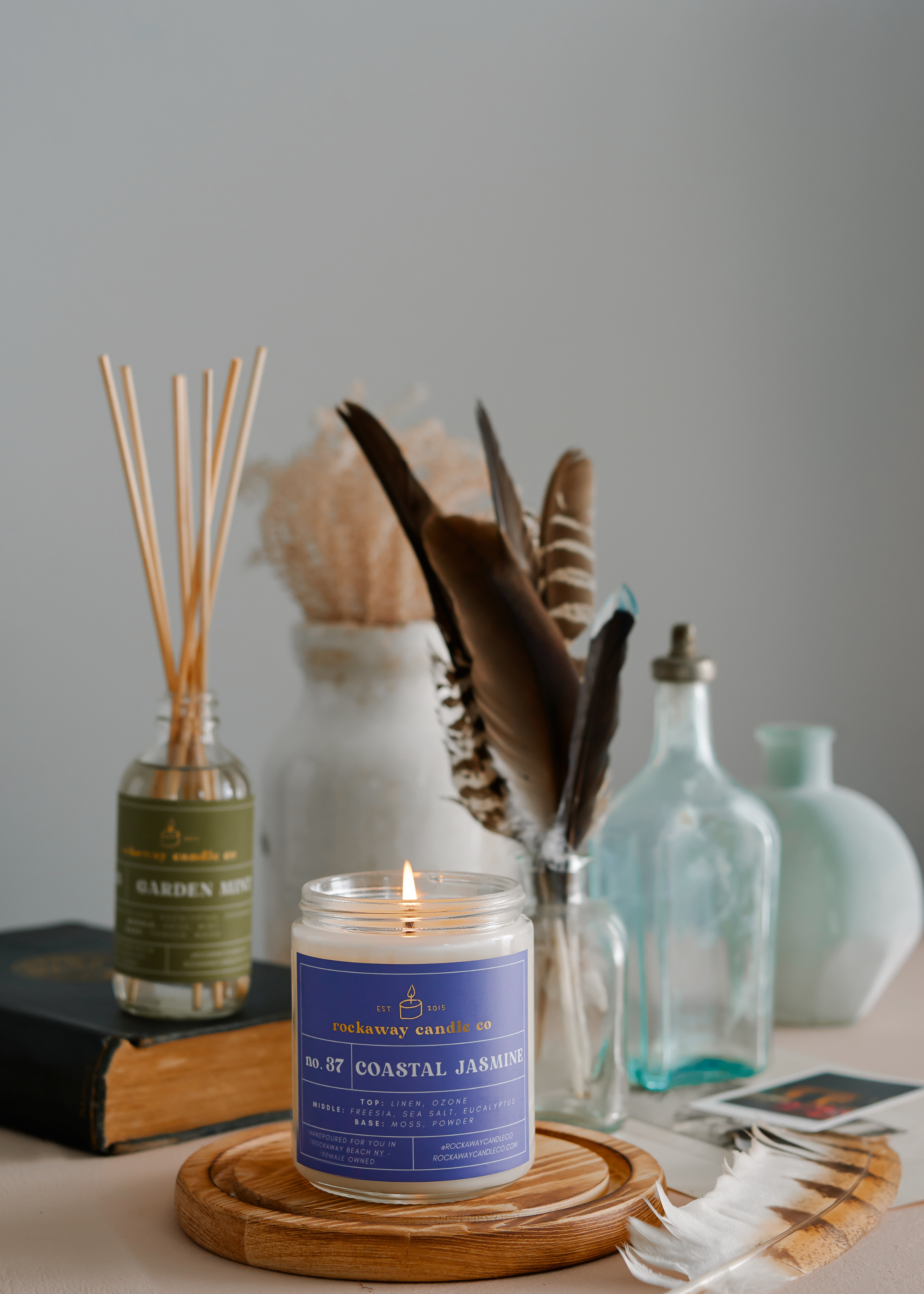 A lit jasmine candle in front of antique blue jars with found feathers and books. Photographed by Philadelphia Product Photographer Tina Crespo