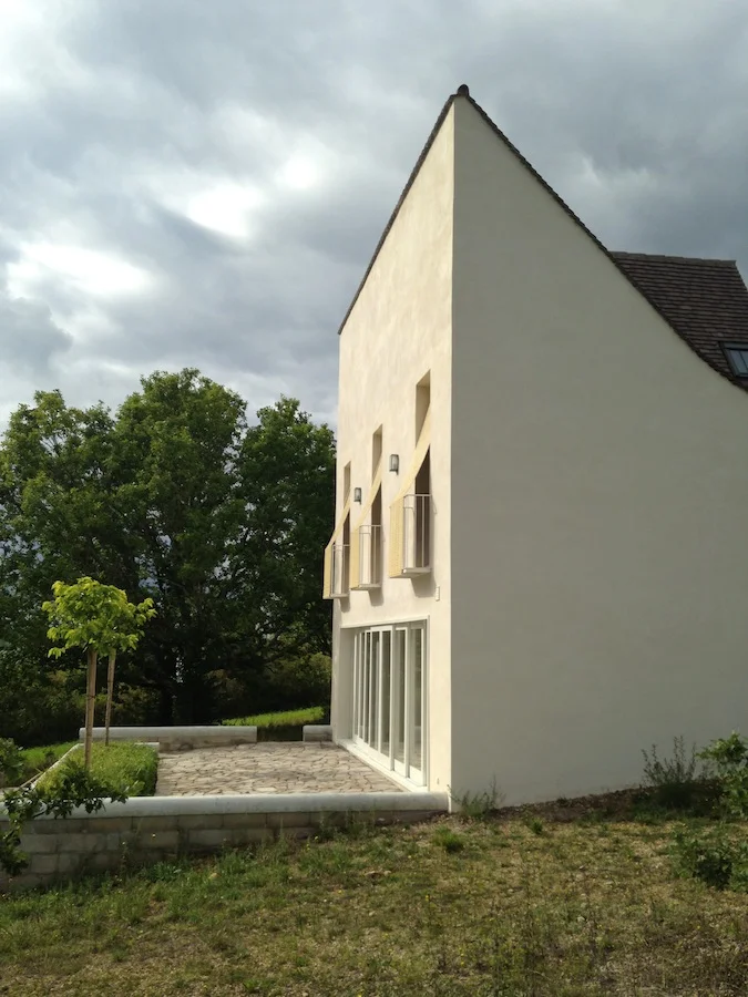 Comment radicaliser des codes bourgeois dans une micro-architecture en site protégé ? Le Roc interroge les codes des architectures moderne, post-moderne et classique pour se recentrer sur son objet, une architecture non convenue, d’une radicalité sans arrogance. Le projet a été conçu avant de trouver le terrain, sur l’hypothèse d’une situation topographique du Causse Lotois, plutôt que sur une parcelle définie. Ce détachement a permis d’aborder un projet autour d’un cahier des charges simple, une table ronde pour 8 personnes, une cheminée, 3 chambres sous rampant, 3 WC, une douche, une baignoire, une buanderie et une piscine. Le terrain est arrivé après la rédaction du projet. Une parcelle en pente de 13 mètres de large, sur 220 mètres de long, avec un dénivelé de 37 mètres. La seule parcelle constructible sur ce flanc de colline. Le projet a donc été posé tout en haut, le plus près possible de la route pour réduire les frais de terrassement et de réseaux. Largeur et longueur de la maison sont définies par la dimension des pierres utilisées pour le rez-de-chaussée. 60x40 cm. Pour compenser la petite surface, les circulations ont été réduites à minima, et deux dispositifs ont été mis en places : une baie de 7,50 mètres en 9 panneaux, et des chambres aux plafonds culminant à 5,35 m avec des portes fenêtres de 2,70 m de haut ouvrant sur des balcons individuels. La maison présente 2 axes de symétrie, un pour la façade sud, l’autre pour la façade nord. La raison provient de la différence de largeur du salon et de la cuisine. La jonction entre ces 2 axes est insoupçonnable puisqu’elle est organisée par un savant dispositif spatial. Le projet n’est pas figé dans sa forme, il peut évoluer, s’étendre. L’arête vive des pignons et la grande façade sud laissent le projet ouvert.