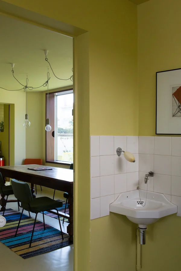  Comment rénover sans nostalgie ? La maison 50 est située dans le centre de Royan, ville bombardée massivement pendant la seconde guerre mondiale, et reconstruite entre 1947 et 1965. La maison, construite par l’architecte parisien Richard Prince en 1952 , est l’ancien syndicat des gens de mer. L’édifice dispose d’un bureau au rez-de-chaussée et d’un logement de fonction à l’étage. Le bâtiment est totalement orienté sur la rue, au nord. Le projet propose deux interventions : il ouvre la maison sur le jardin, et crée une porosité transversale. Les pièces d’origines sont conservées dans leur forme originelle, le programme s’adapte. L’intention architecturale du projet initial est conservée, mais la rénovation rectifie certains inachèvements. Un travail précis de préservation / remplacement / création est mené. Les portes intérieures, les poignées, les menuiseries extérieures sont nettoyées et réparées. Les menuiseries en niangion au mécanisme d’époque sont révisées. De nouveaux volets en pin d’orégon sont fabriqués. Les sols existants sont tous conservés, avec l’ajout de «rustines» aux endroits où le sol est abîmé. Deux portes intérieures sont créées, pour compléter les porosités transversale et longitudinale du projet. Chaque pièce compte alors entre deux et trois portes ; la circulation dans la maison devient fluide. Côté jardin, deux grandes baies en aluminium, fixées en applique, constituent la seule intervention actuelle. Le caractère exceptionnel des deux baies rend hommage à l’enthousiasme de la reconstruction, au goût du défi. Il prolonge l’histoire de la maison, et l’accroche au XIX ème siècle, sans nostalgie. L’intérieur est abordé avec le même souhait. Pour résister à la tentation d’une approche décorative d’un esprit 50 revisité, la maison est conçue comme un monochrome jaune/vert. Trois nuances sont réparties en fonction de l’étage, du programme, et de l’orientation de la maison, selon une approche scientifique (optique) de la couleur. Toute référence au blanc est évacuée. Le jaune devient un nouveau blanc et donne l’illusion d’habiter un tableau. 