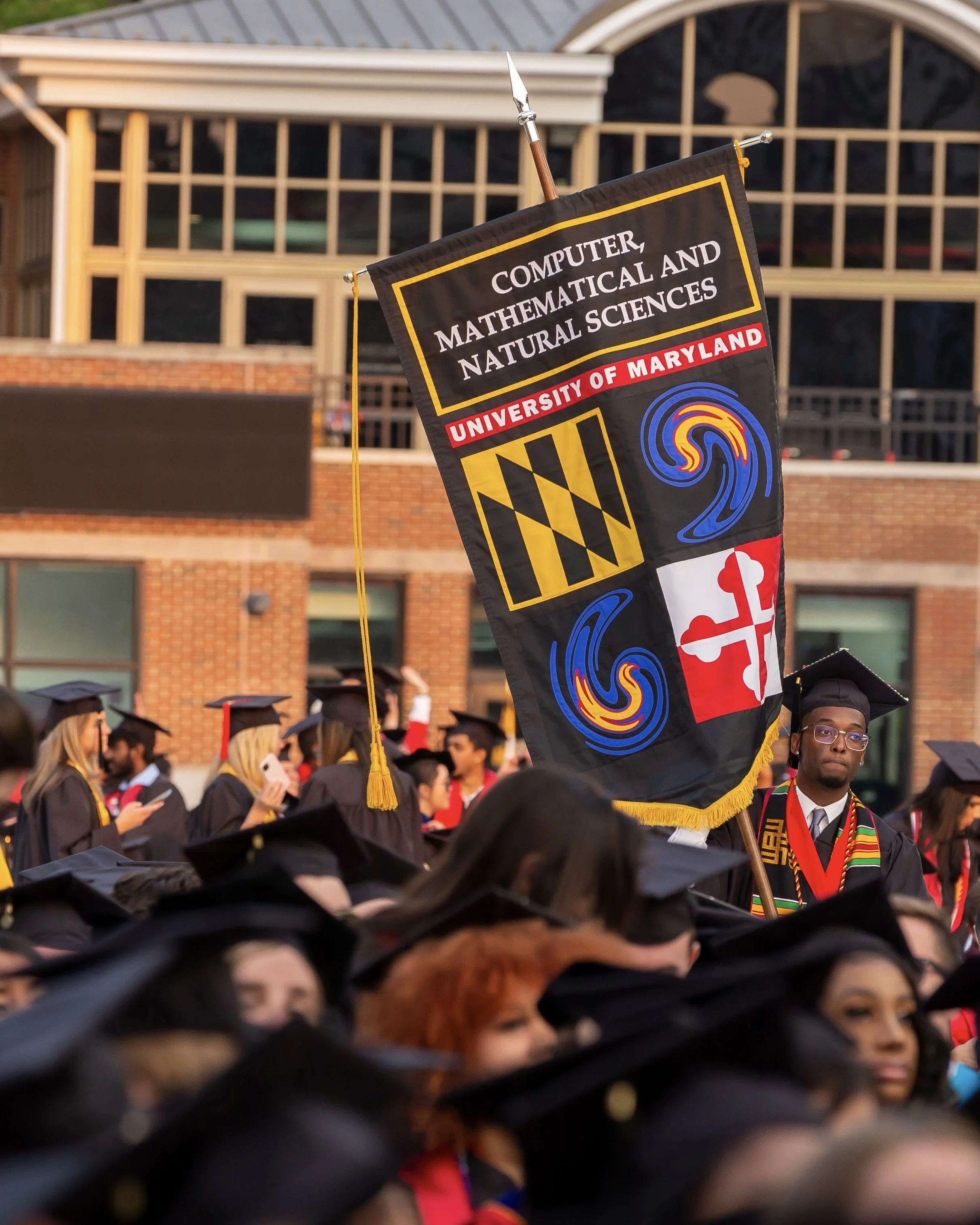 UMD graduate carries a banner for the College of CMNS amongst a sea of fellow graduates.
