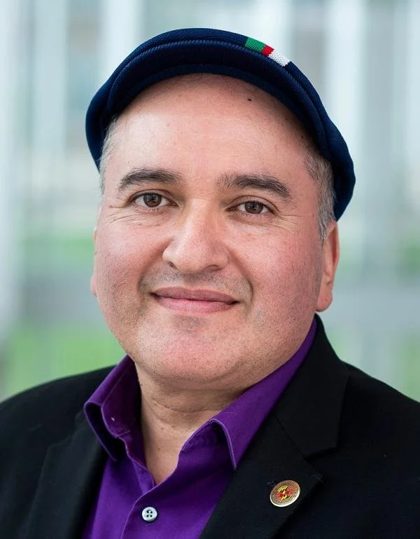 Portrait of a man in a purple shirt, hat, and black blazer.