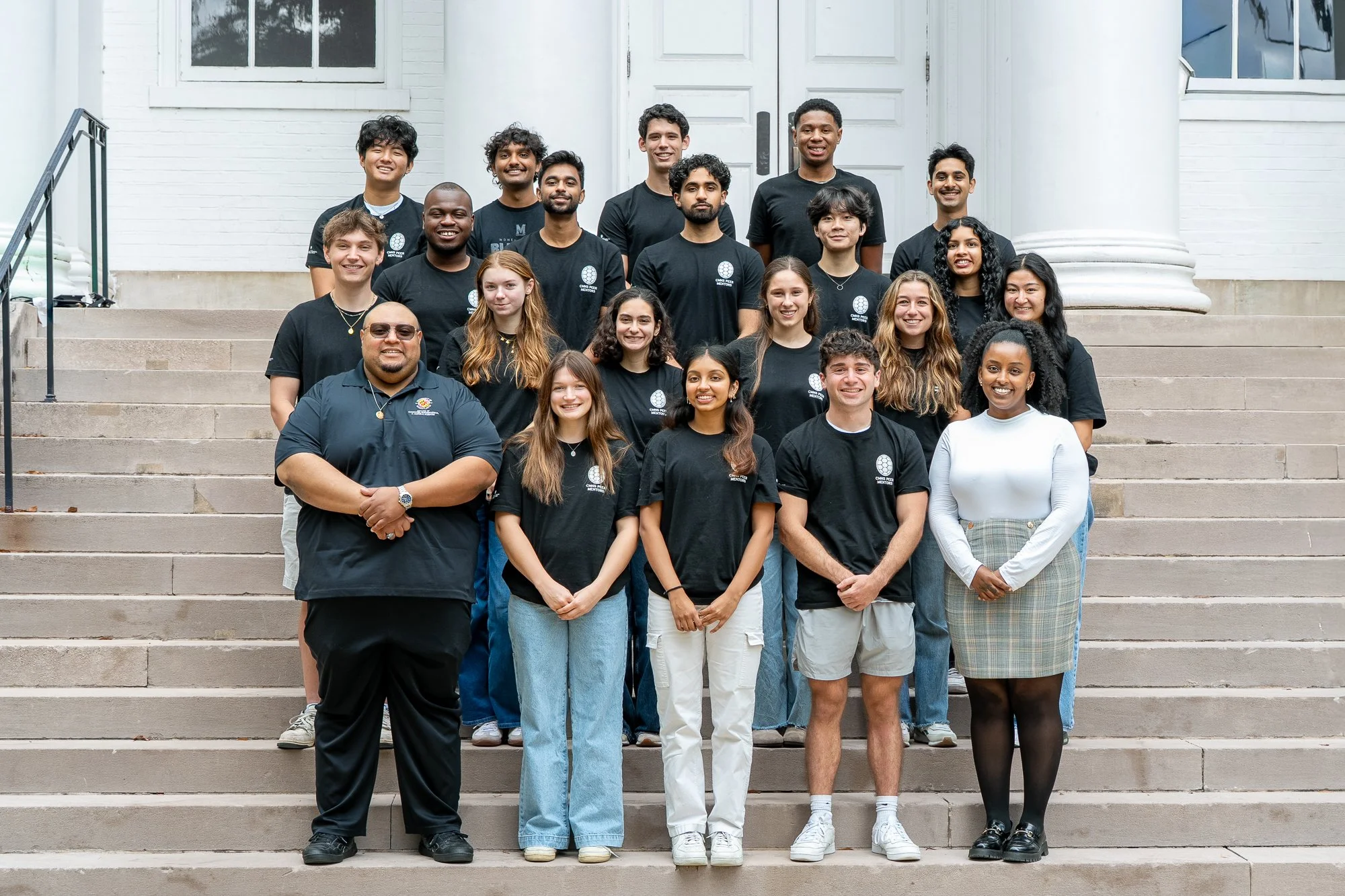 Group of college students in black t-shirts standing on the stone staircase of a white building.