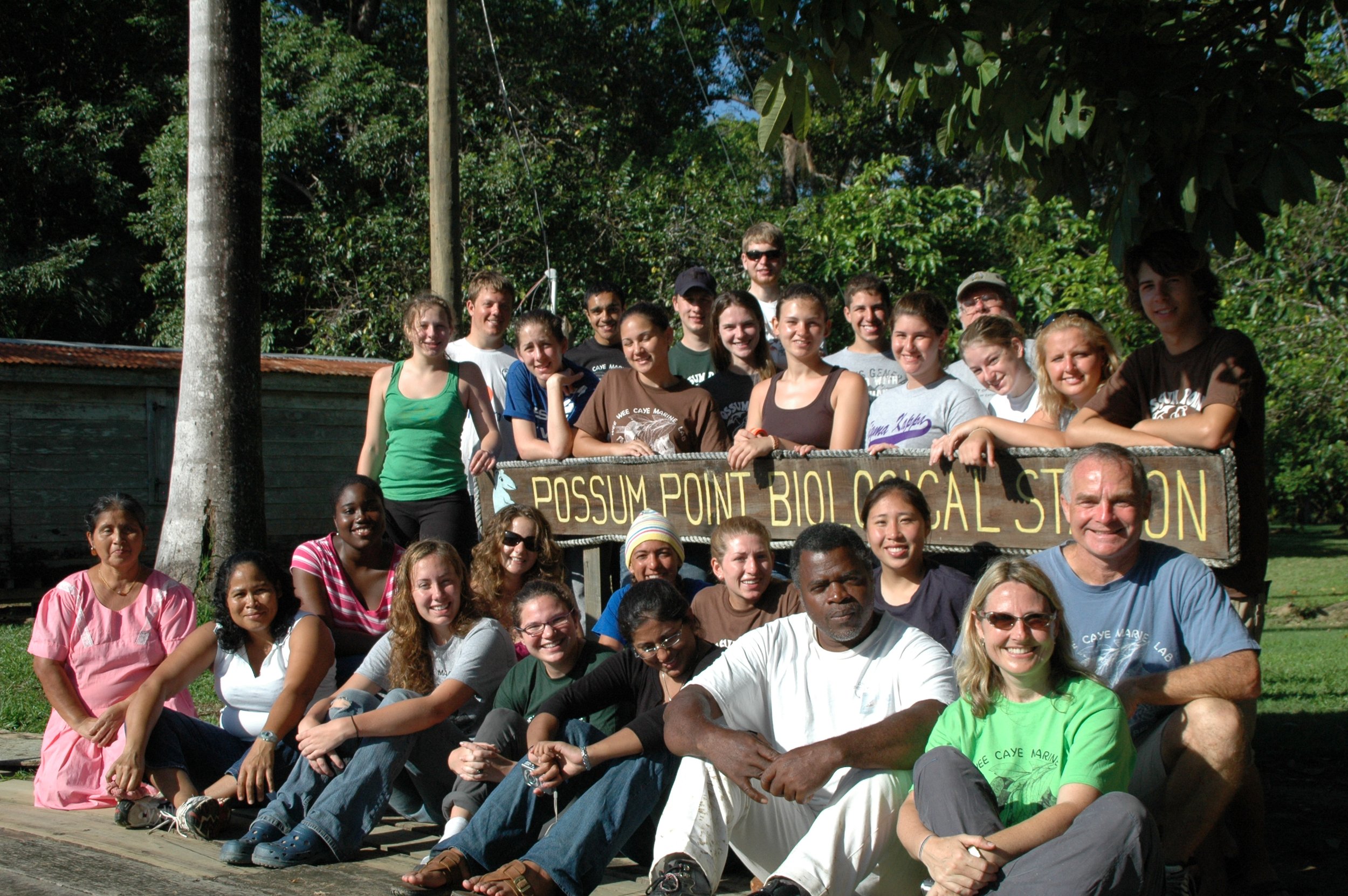 A group of people in a tropical environment standing amidst a wooden sign that reads "Possum Point Biological Station."