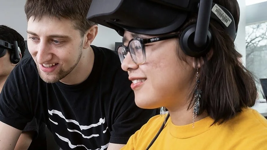 Young woman in yellow shirt and glasses looks at a computer screen next to a young man in a black shirt.