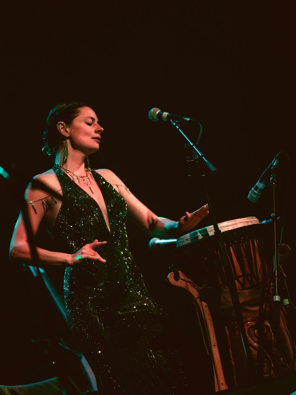 Bonnie Paine of Elephant Revival performing live on stage, playing djembe drum in a sequined dress.