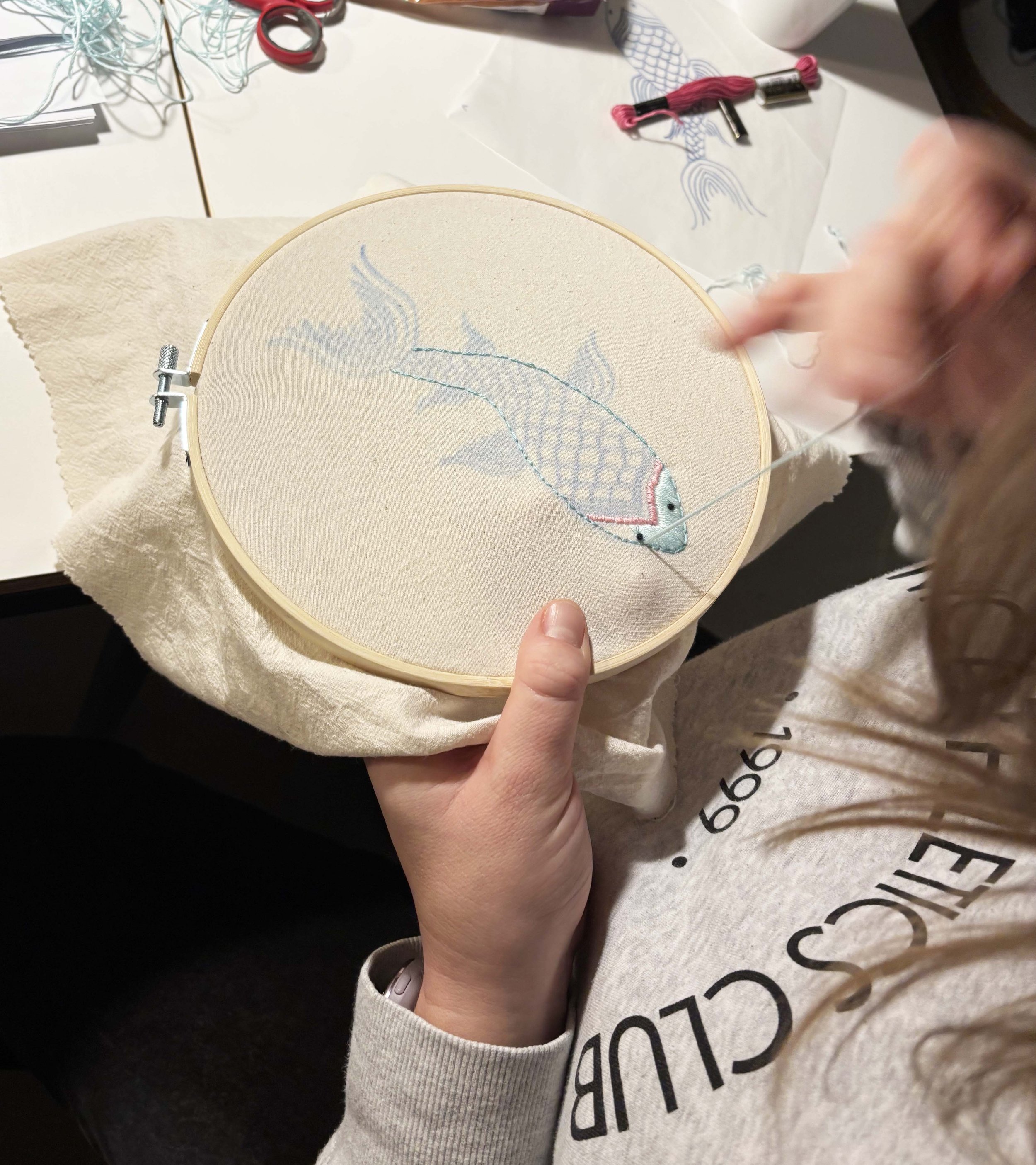 Eine Person stickt ein Fischdesign auf ein kreisförmiges Stoffstück in einem Stickrahmen. Es sind Stickgarn und andere Stickmaterialien auf dem Tisch zu sehen.
