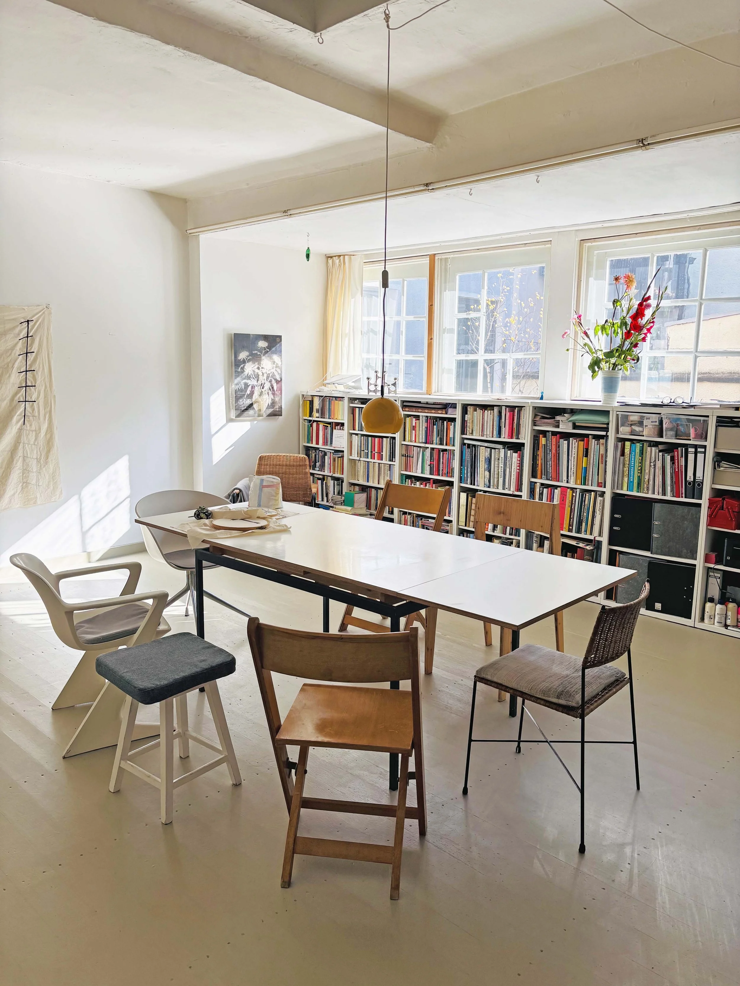 Heller Raum mit Tisch, bunten Stühlen, Bücherregal und großen Fenstern mit Blick nach draußen, Sonnenlicht fällt auf den Boden.
