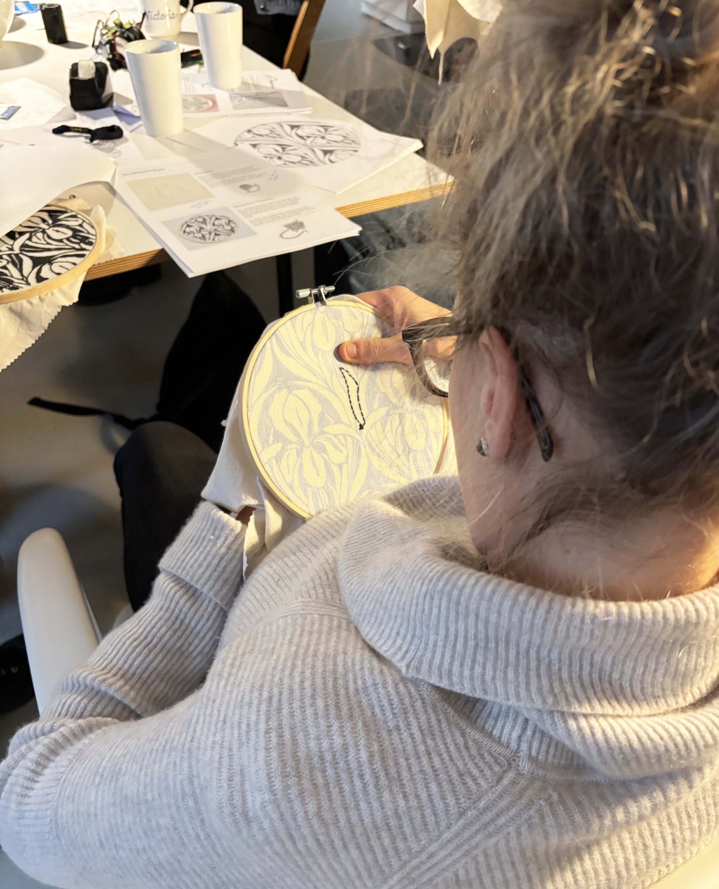 Eine Person, wahrscheinlich eine ältere Frau, näht an einem Stickrahmen mit einem gelb gemusterten Stoff. Der Tisch vor ihr ist mit verschiedenen Papieren, Stickmustern und anderen Nähutensilien bedeckt, und im Hintergrund sind weiße Tassen sowie wei