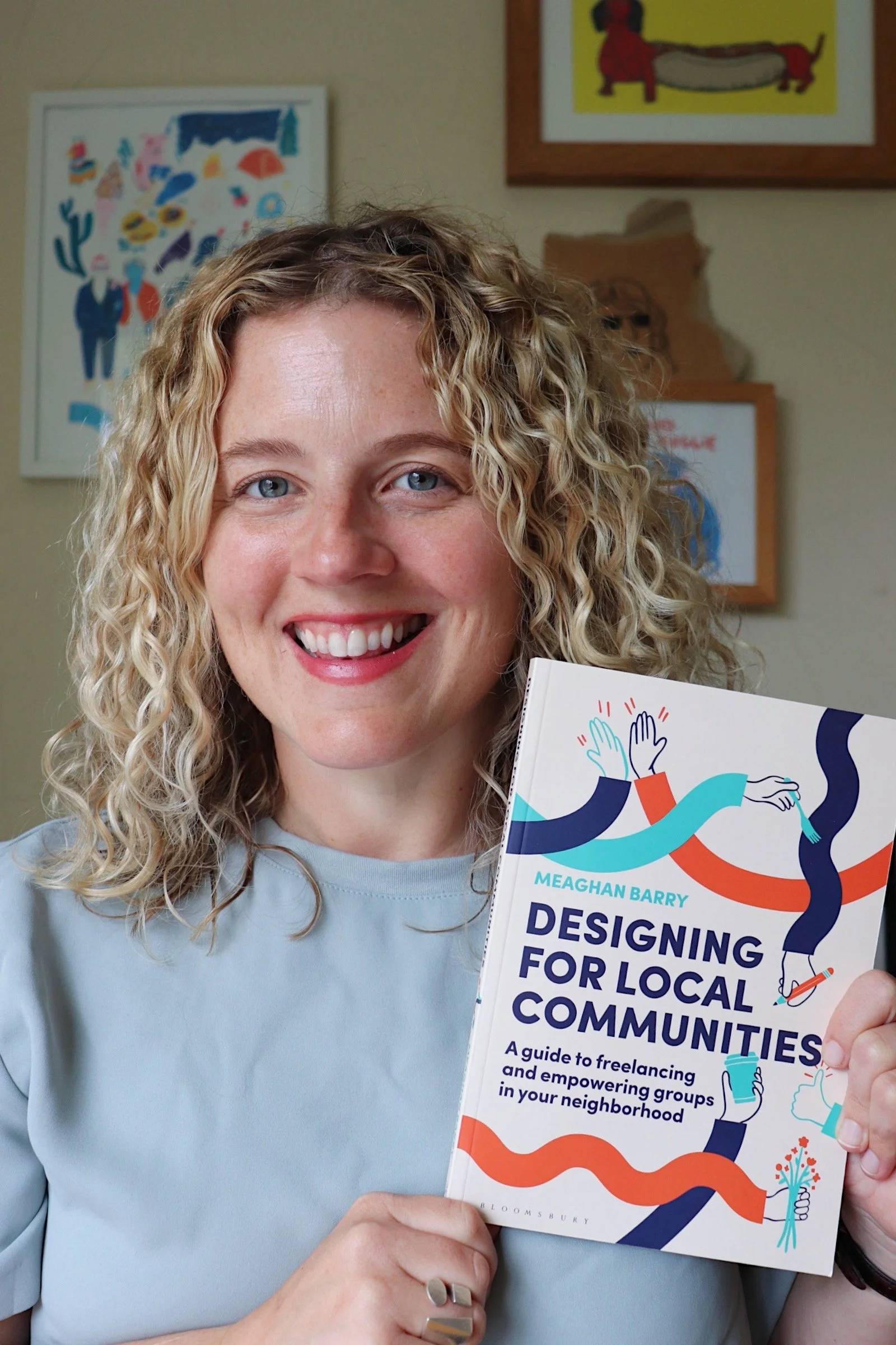 A woman with curly blonde hair smiling and holding a book titled 'Designing for Local Communities' by Meaghan Barry. The background features framed artwork on a wall.