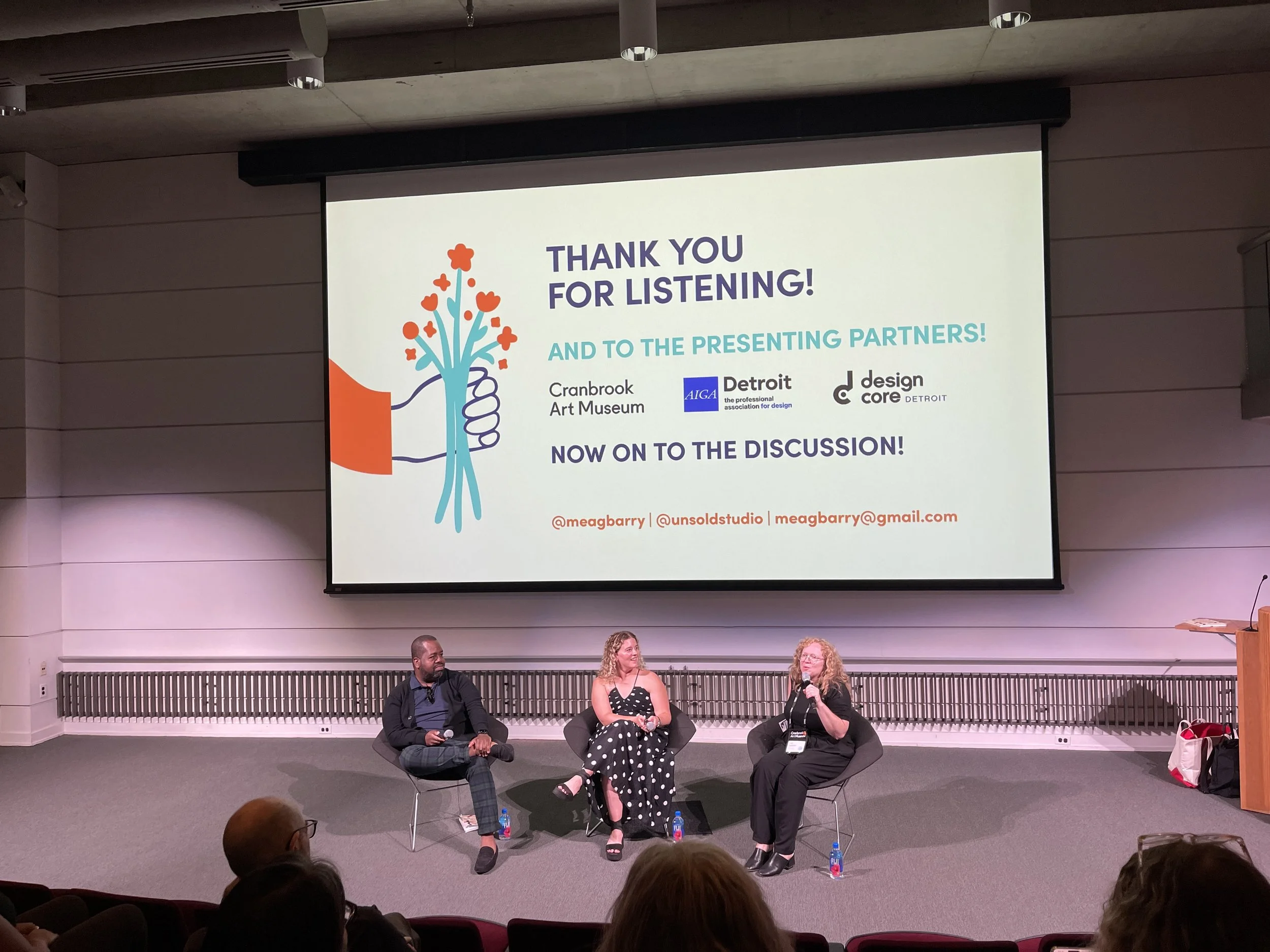 A panel discussion is taking place in an auditorium with three speakers seated on stage in front of a large screen. The screen displays a thank you message to the audience, logos of sponsors, and contact information. The auditorium has an audience seated facing the stage.