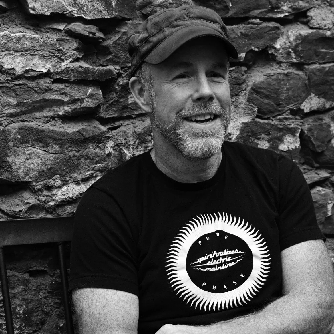 black and white photo of James Belflower in a black hat, black shirt with a Spiritualized logo on it.