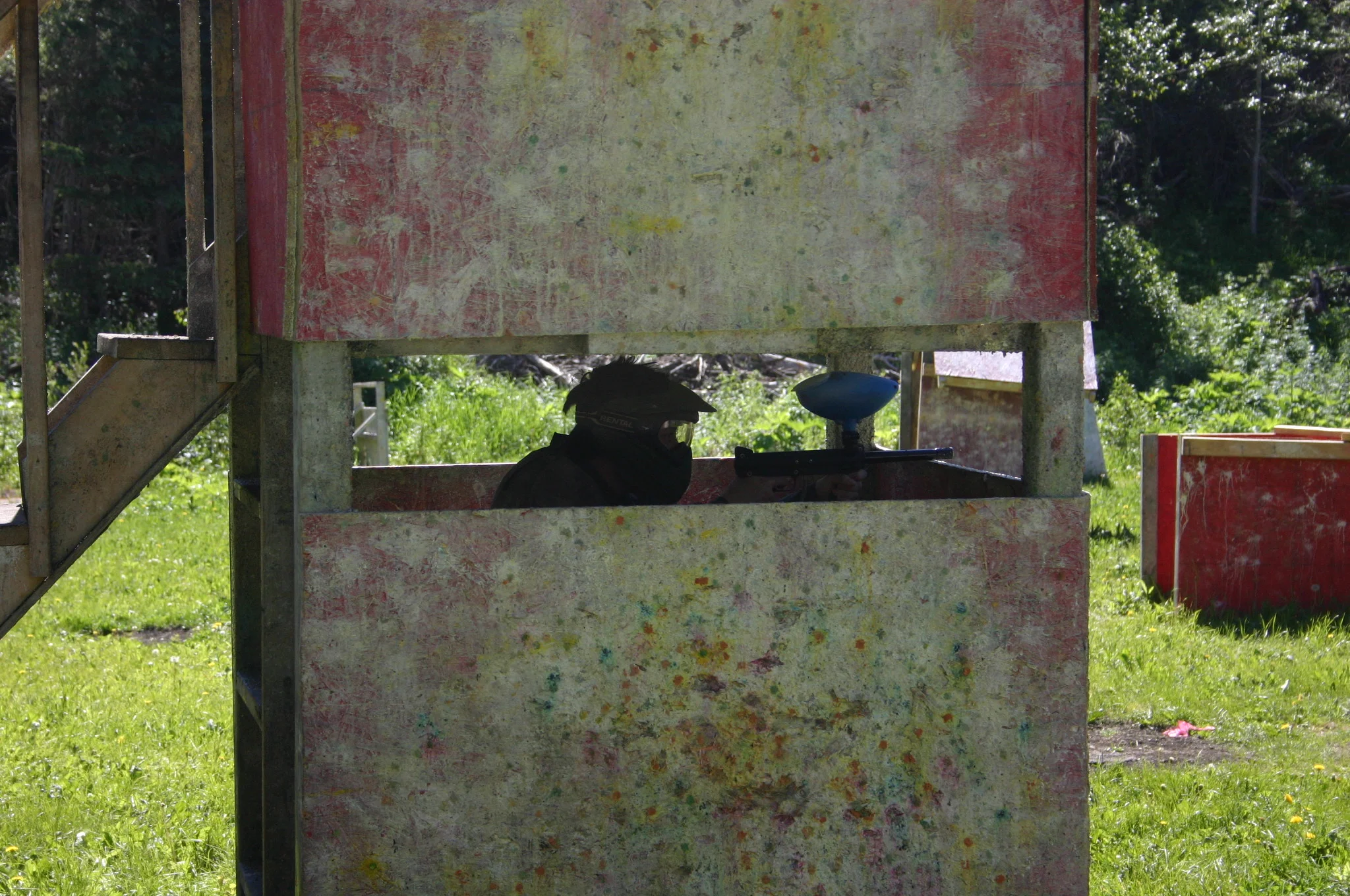 Person playing paintball behind a wooden bunker, holding a paintball marker.