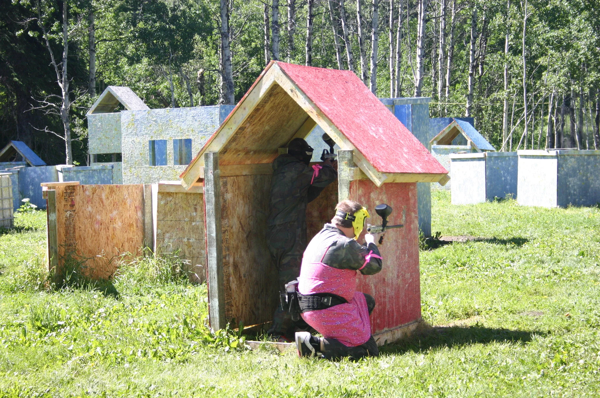 Two people playing paintball, one kneeling and one standing inside a small wooden shelter with a red roof, in an outdoor field with wooden structures and trees in the background.