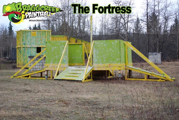 Paintball arena structure labeled 'The Fortress' from Bragg Creek Paintball, featuring yellow and green wooden ramps and walls in a wooded area.