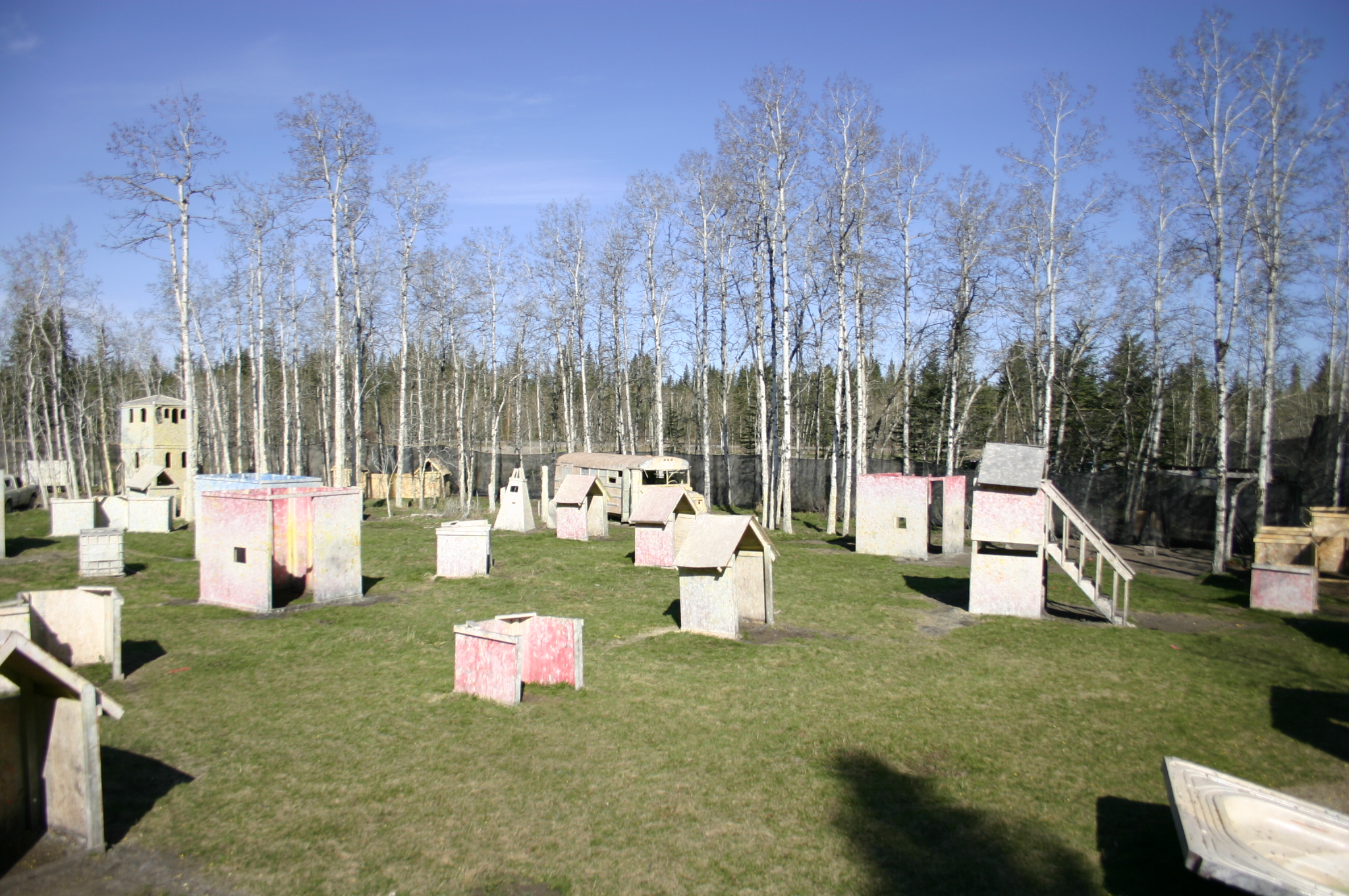 Outdoor paintball field with various bunkers and obstacles set up on grassy terrain, surrounded by leafless trees against a clear sky.