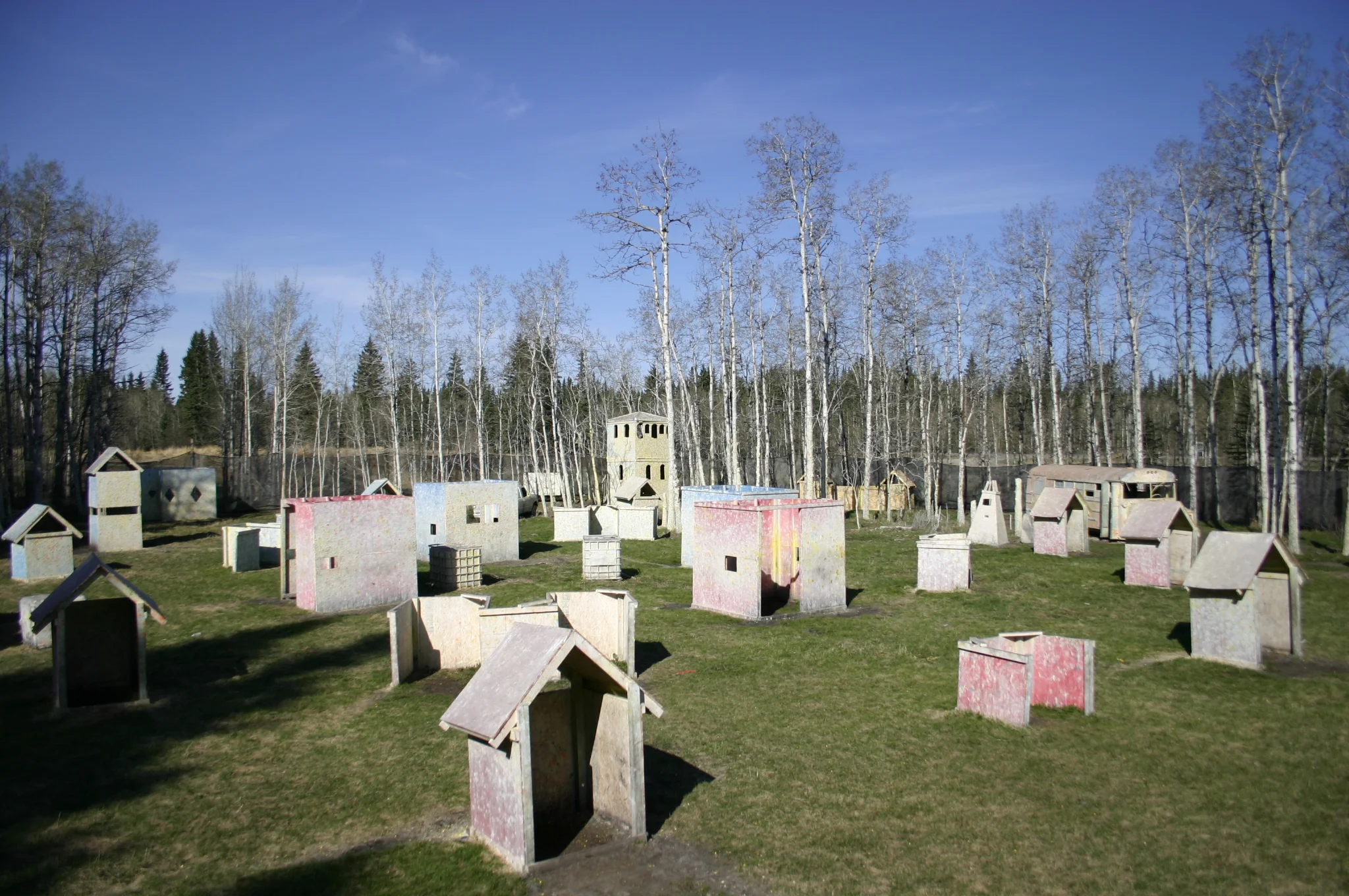 Outdoor paintball field with wooden bunkers and structures in a grassy area surrounded by trees.