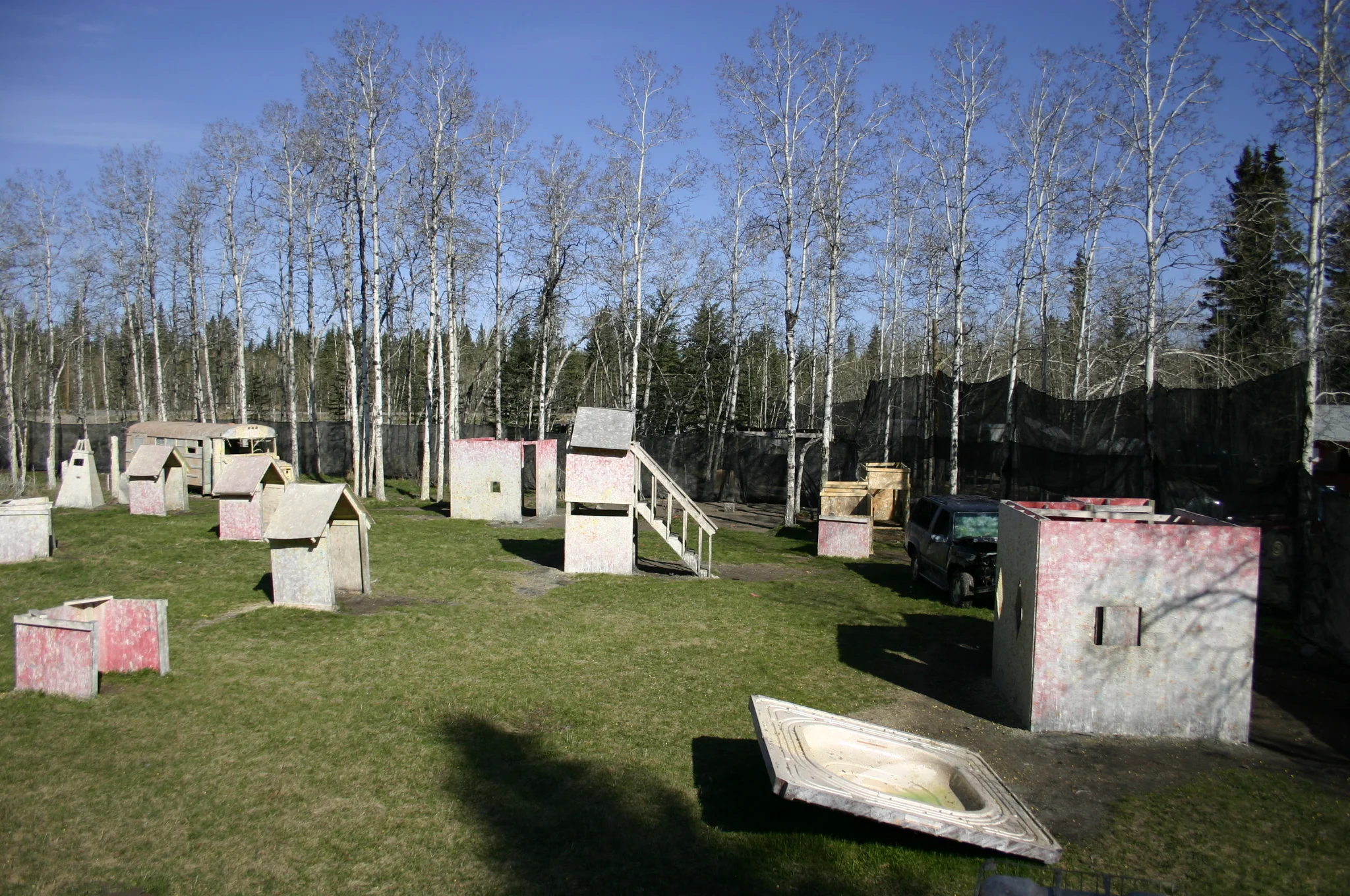 Outdoor paintball field with various wooden structures and obstacles, surrounded by trees and a fence.