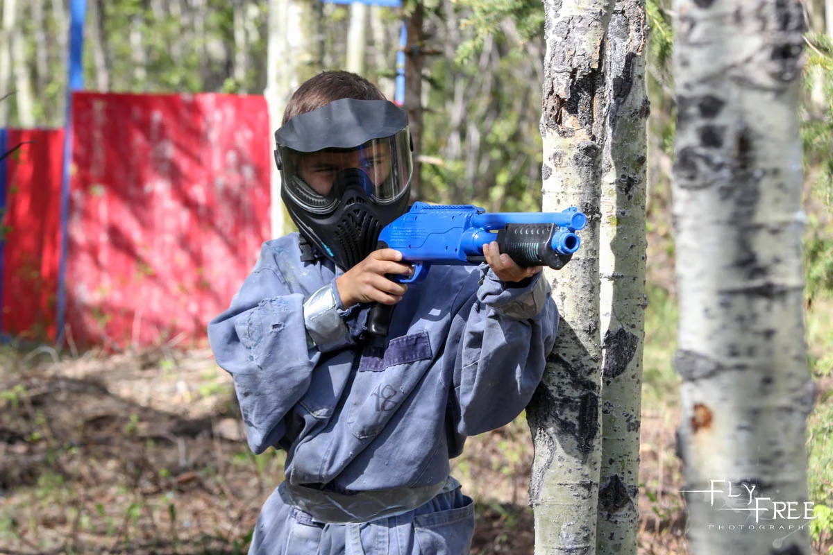 A young boy wearing a paintball mask and gray coveralls, aiming a blue paintball gun behind a tree in a forested area, with colorful barriers in the background.