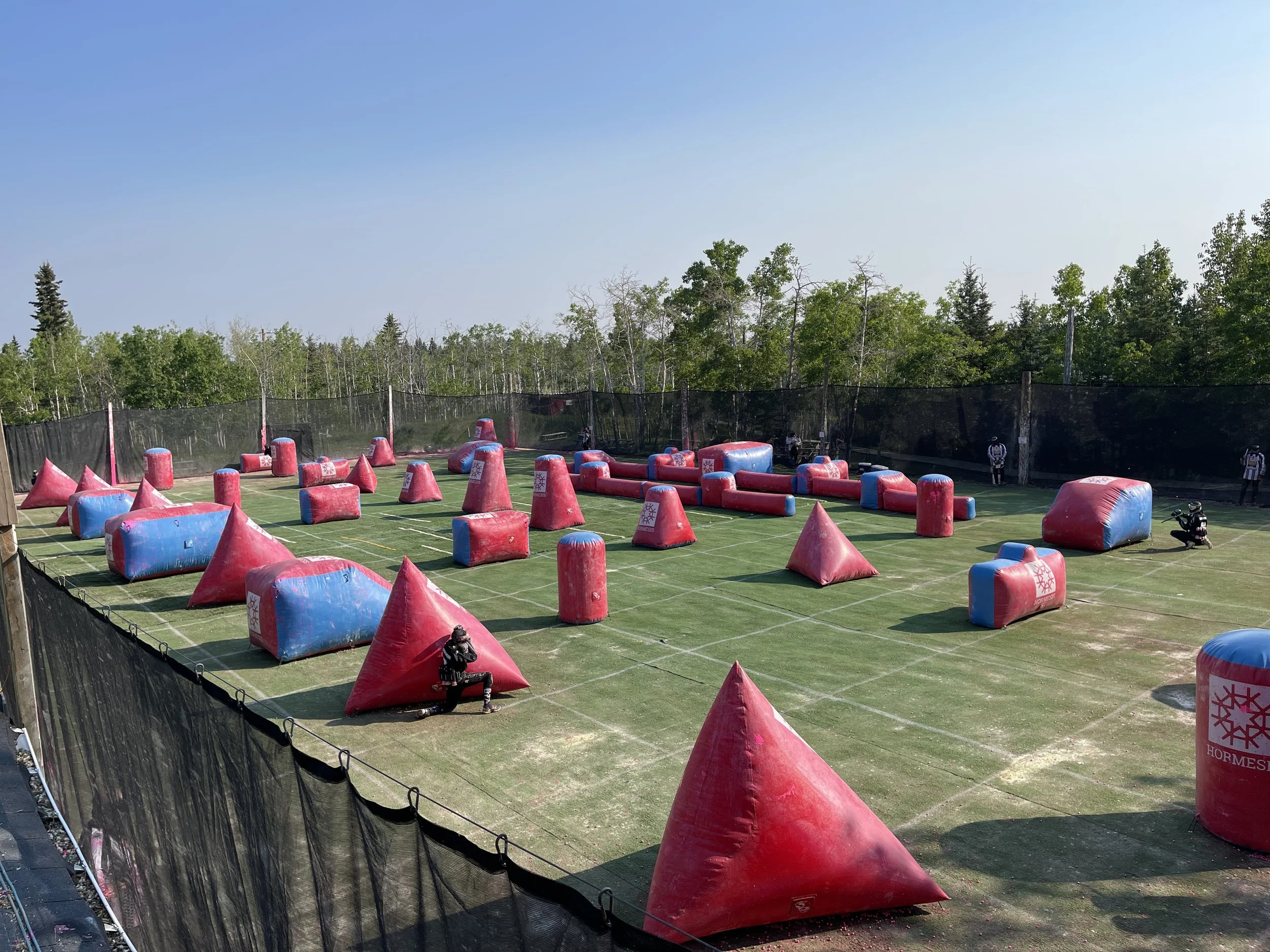 An outdoor paintball field with inflatable obstacles in red and blue, surrounded by a black net, with trees in the background and players wearing protective gear.