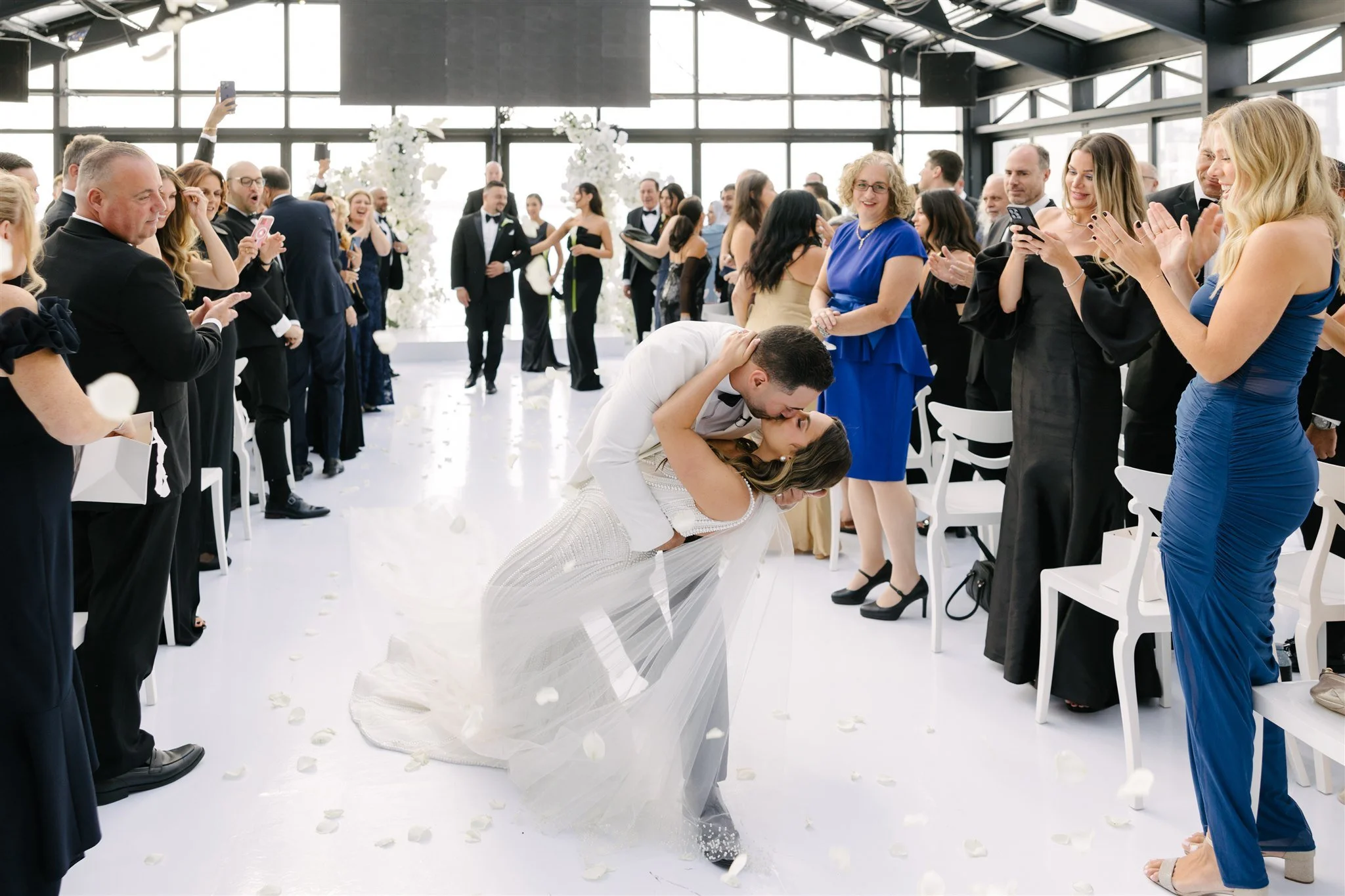Bride and groom sharing a kiss while bending down, guests clapping and taking photos at a wedding reception in a bright, modern venue.