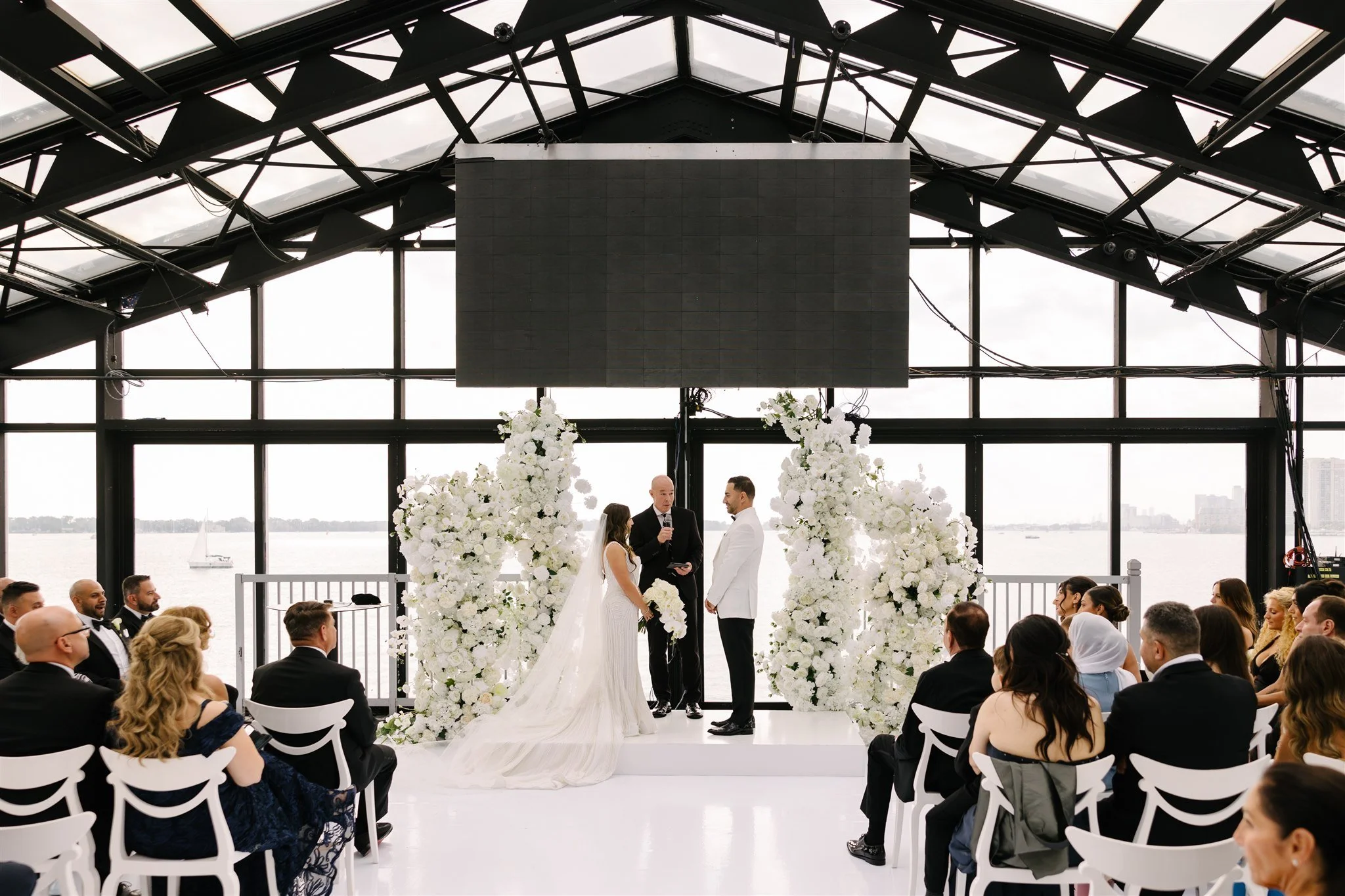 Wedding ceremony in a glass-walled venue with a bride and groom standing before an officiant, surrounded by large white floral arrangements, with guests seated watching. Water and sailboats visible through the windows.