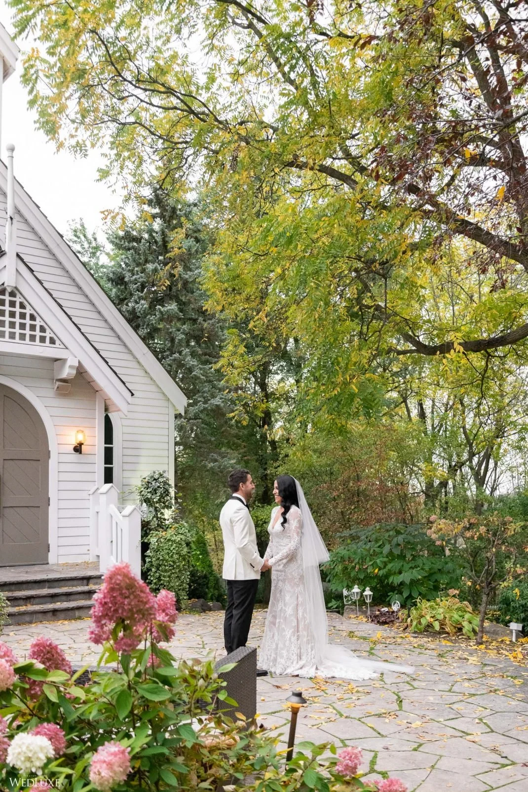 A bride and groom holding hands and smiling at each other outside near a white house with a pathway and surrounded by greenery and pink hydrangeas.