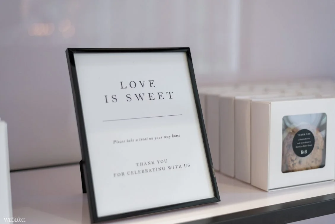 A framed sign on a table that says "Love is Sweet" with additional text about taking a treat home and thanking guests for celebrating.